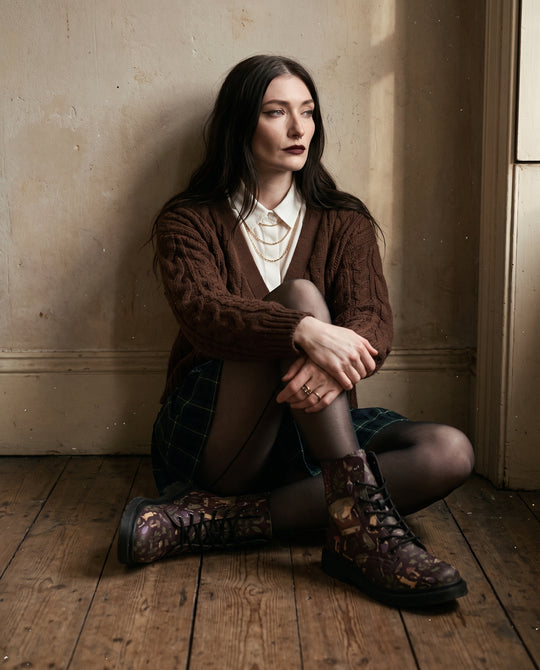 Woman sitting on a wooden floor wearing Toad Crypt Boots with a brown cable cardigan and tartan skirt